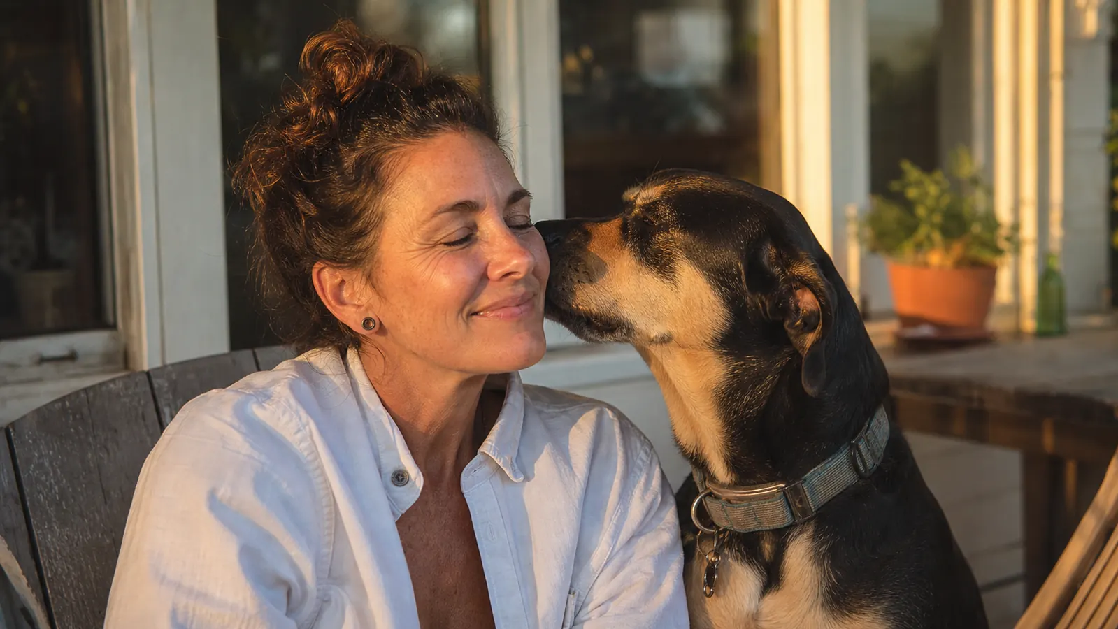 Woman and her dog sitting together on a back porch, the dog nuzzling close to her face, both relaxed and happy
